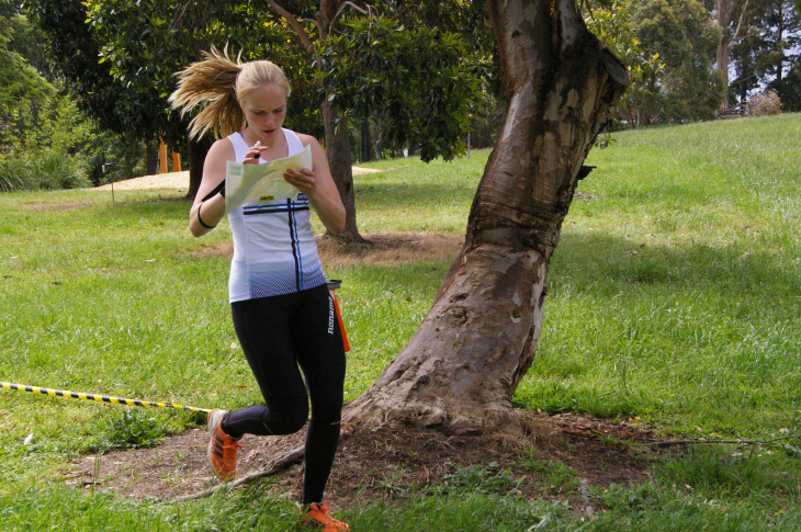 Female runner in a white sleeveless top reads a map while moving along a grassy park trail, with a yellow-and-black taped boundary in the foreground.